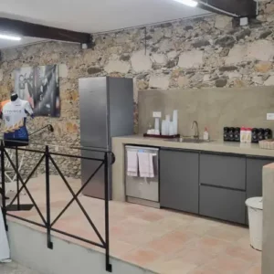 Cuisine rustique moderne avec mur en pierre apparente, électroménagers inox (frigo, lave-vaisselle) et comptoir gris. Un maillot CYNISCA est exposé.