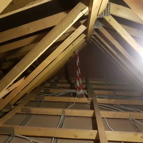 Structure de charpente en bois sous un toit, avec des solives, un marquage rouge et blanc suspendu, et des câbles électriques visibles.