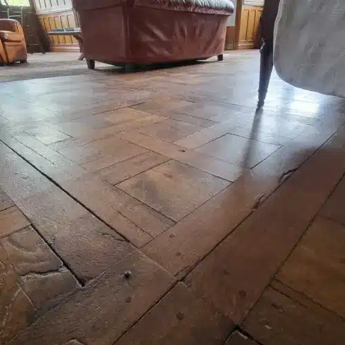 Parquet ancien en bois foncé avec motif à chevrons et larges lames, éclairé par la lumière du jour près d'un canapé en cuir marron.