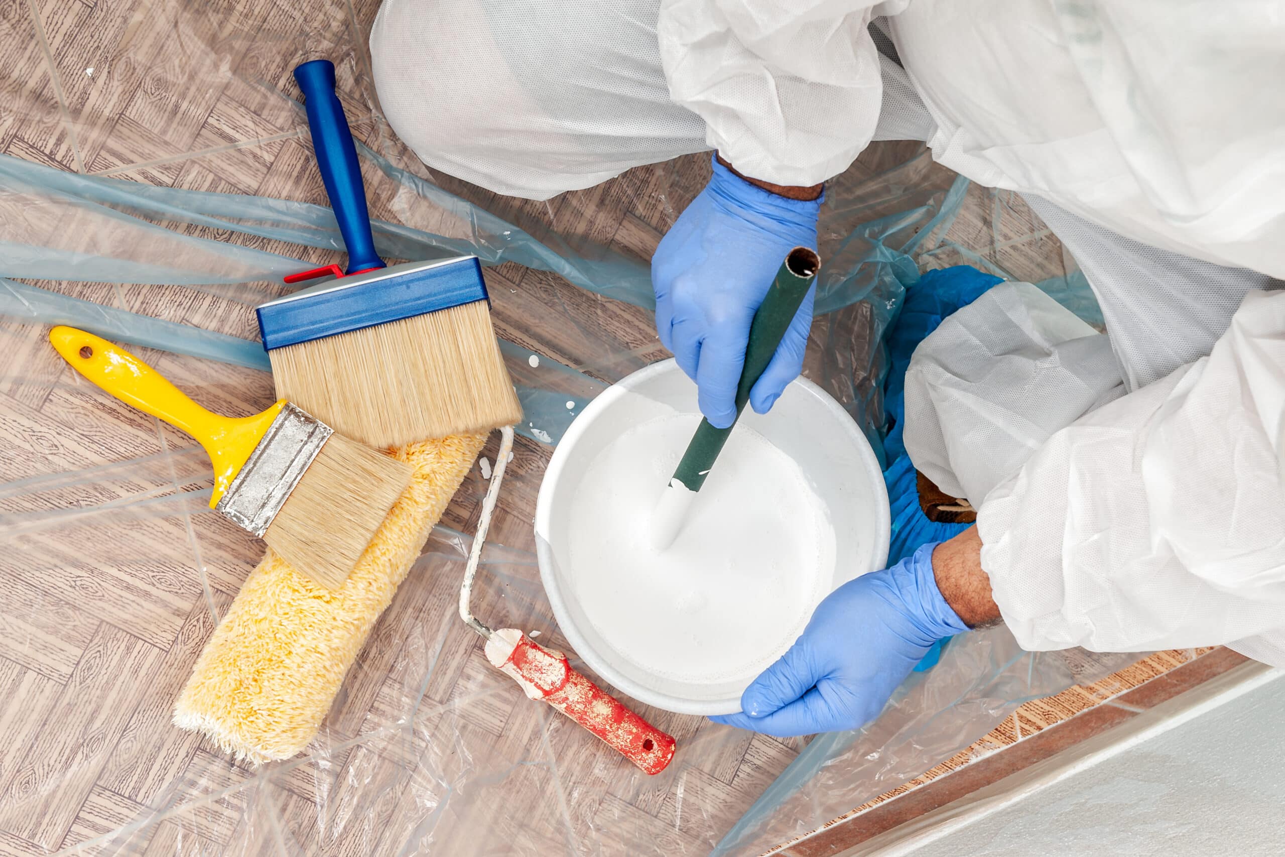 Peintre en combinaison de protection, ganté, mélange de peinture blanche dans un seau avec un bâton, entouré d'outils.